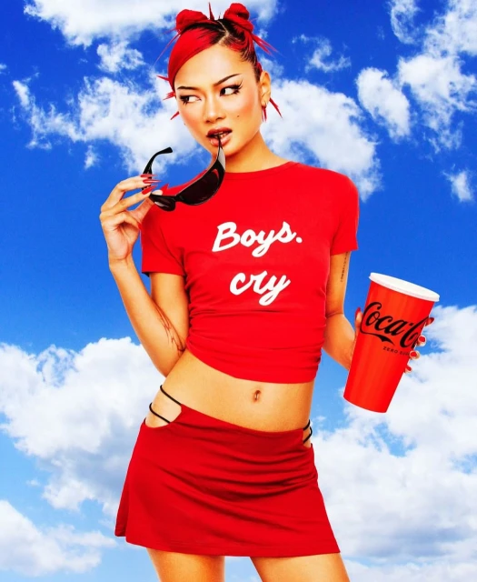 Young woman with red hair, wearing a red t-shirt and skirt, holds sunglasses and a Coca-Cola cup against a cloudy sky background.