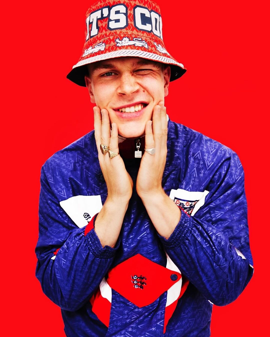 A young man in a patterned bucket hat and retro jacket smiles and winks, holding his hands to his face against a solid red background.