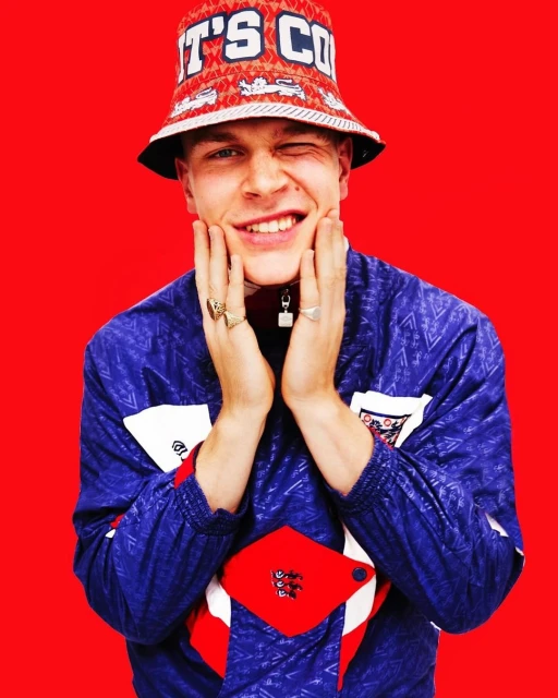 A young man in a patterned bucket hat and retro jacket smiles and winks, holding his hands to his face against a solid red background.