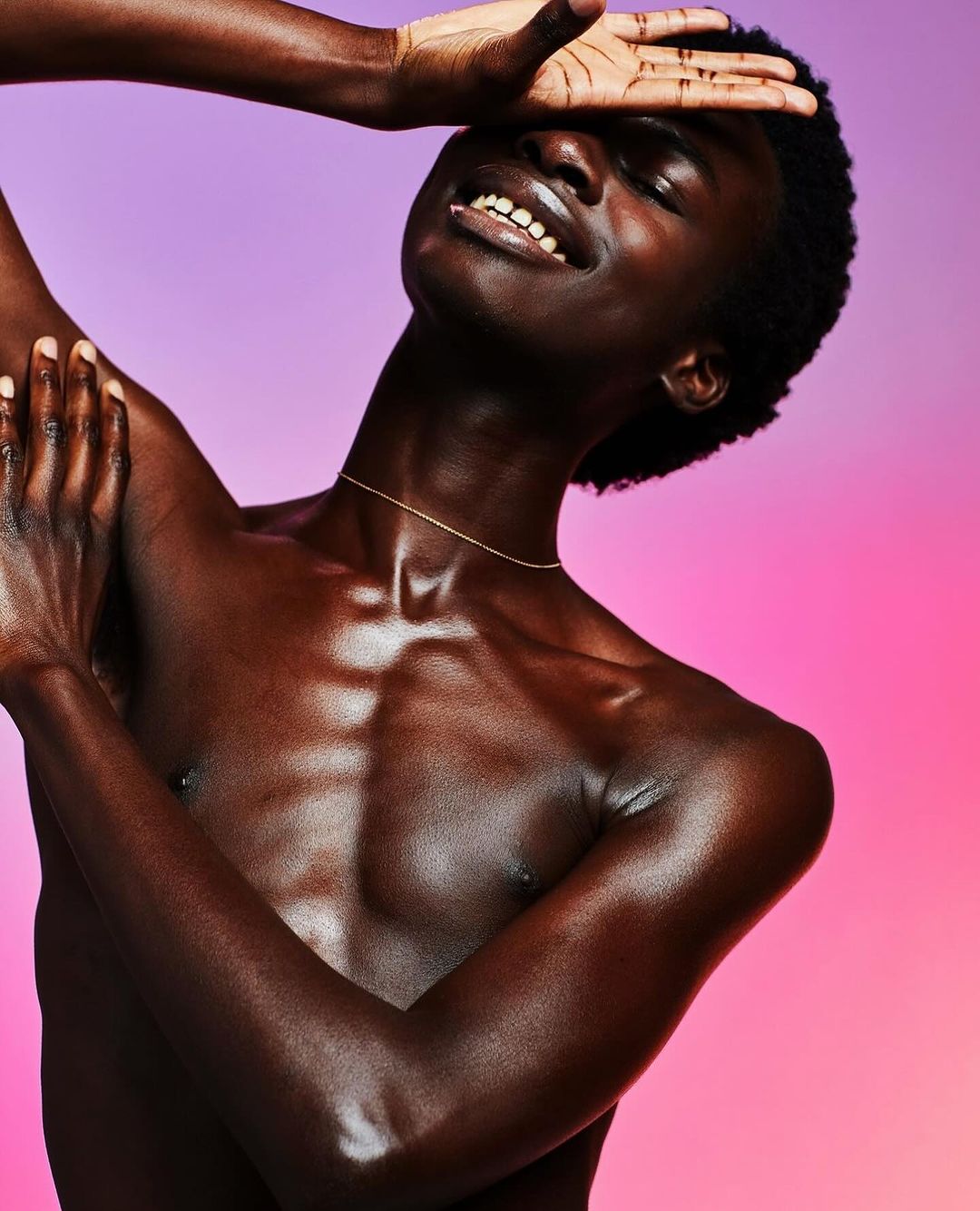 Portrait of an adult male with dark skin, bare chest and a slight smile posing in front of a pink and purple gradient background.