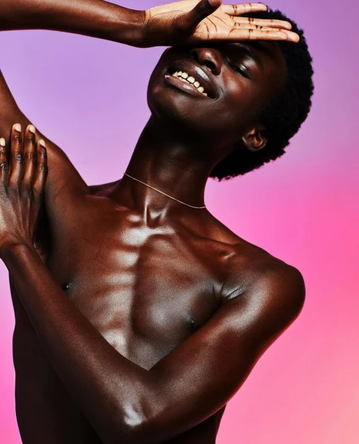 Portrait of an adult male with dark skin, bare chest and a slight smile posing in front of a pink and purple gradient background.