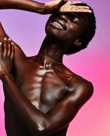 Portrait of an adult male with dark skin, bare chest and a slight smile posing in front of a pink and purple gradient background.