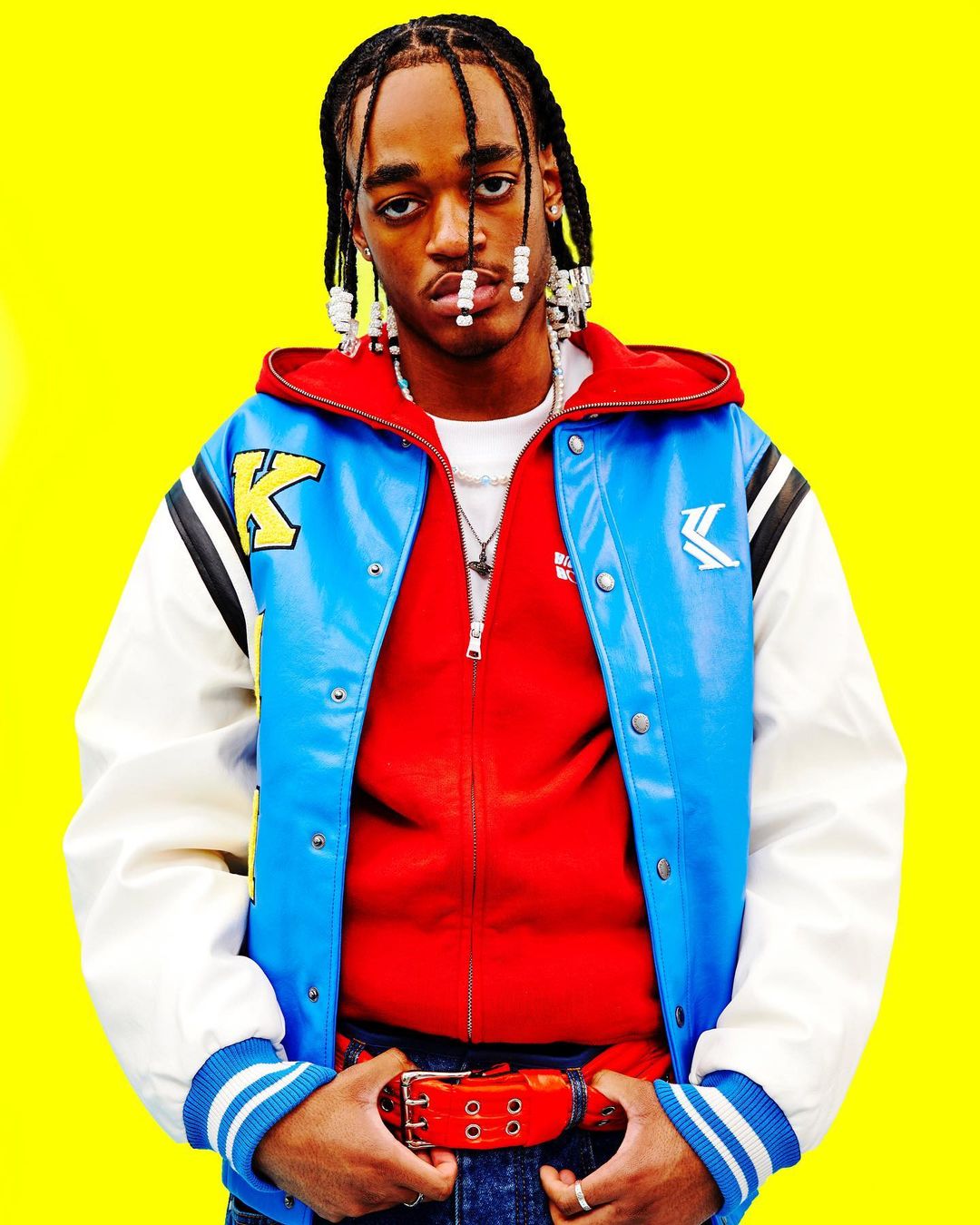 Portrait of a young adult male with beaded braids wearing a blue and white varsity jacket against a bright yellow background.