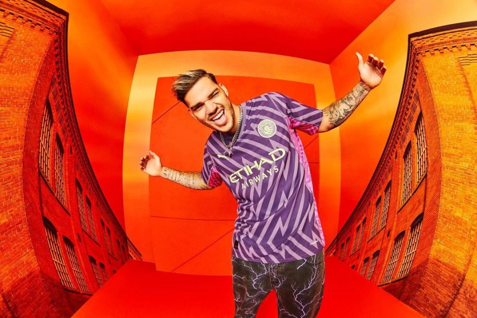 Man in soccer jersey poses in studio with red backdrop and brick wall detail, expressive pose, tattoos, and streetwear style.