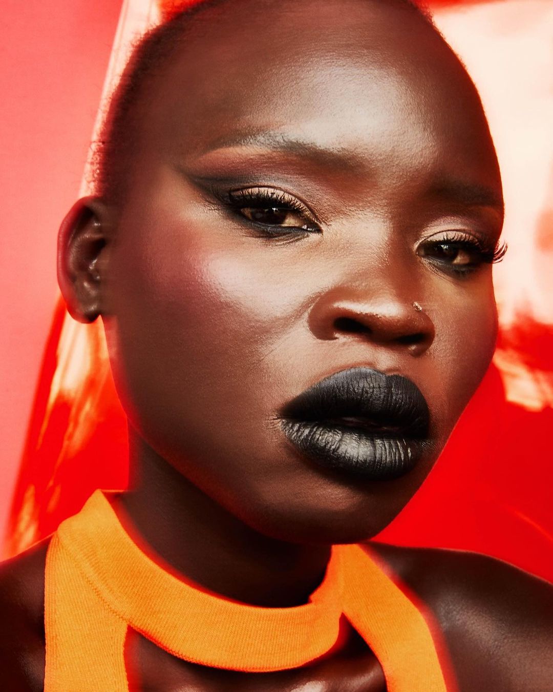 Close-up portrait of an African American adult female with short hair, bold eye makeup, black lipstick, wearing an orange top on a red background.