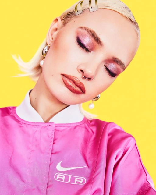 A young adult woman with blonde hair and pink makeup wears a pink Nike jacket against a bright yellow background in a fashion portrait.