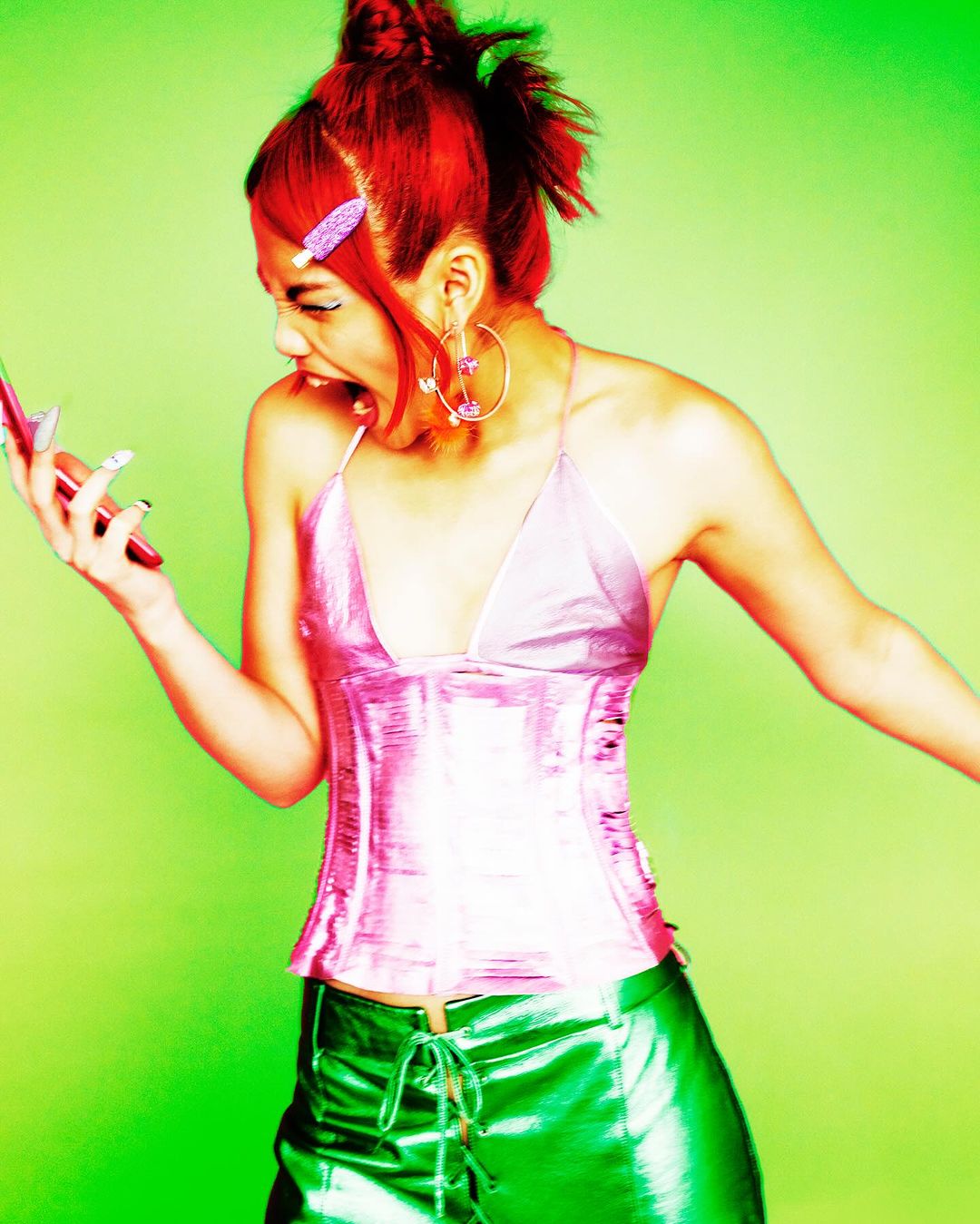 A young woman with bright red hair wearing shiny, futuristic style clothing is yelling while holding a cell phone in her hand.