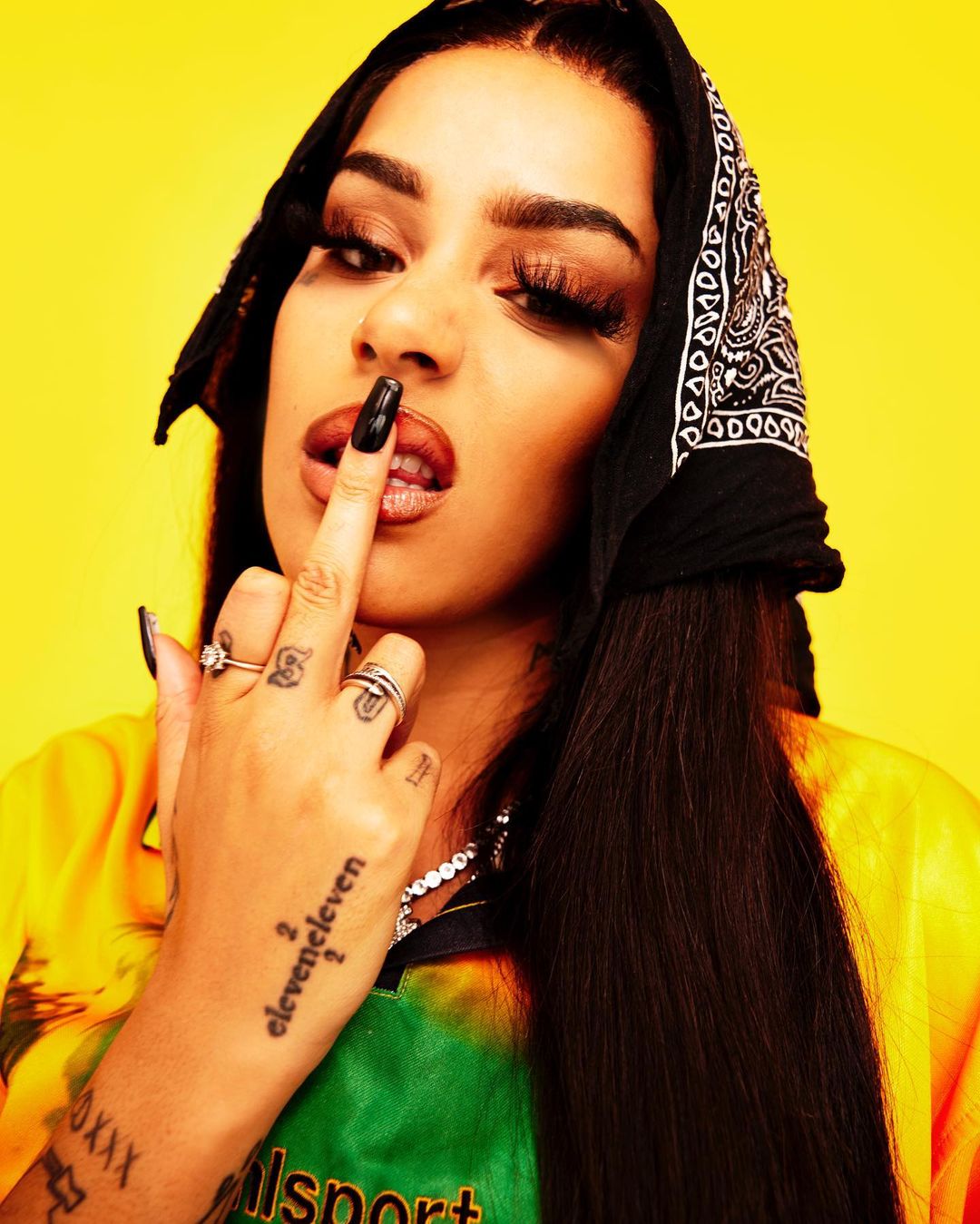 Close-up shot of a woman wearing a bandana with dark hair, black nails, and jewelry, posing against a vibrant yellow backdrop.