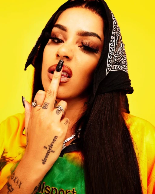 Close-up shot of a woman wearing a bandana with dark hair, black nails, and jewelry, posing against a vibrant yellow backdrop.