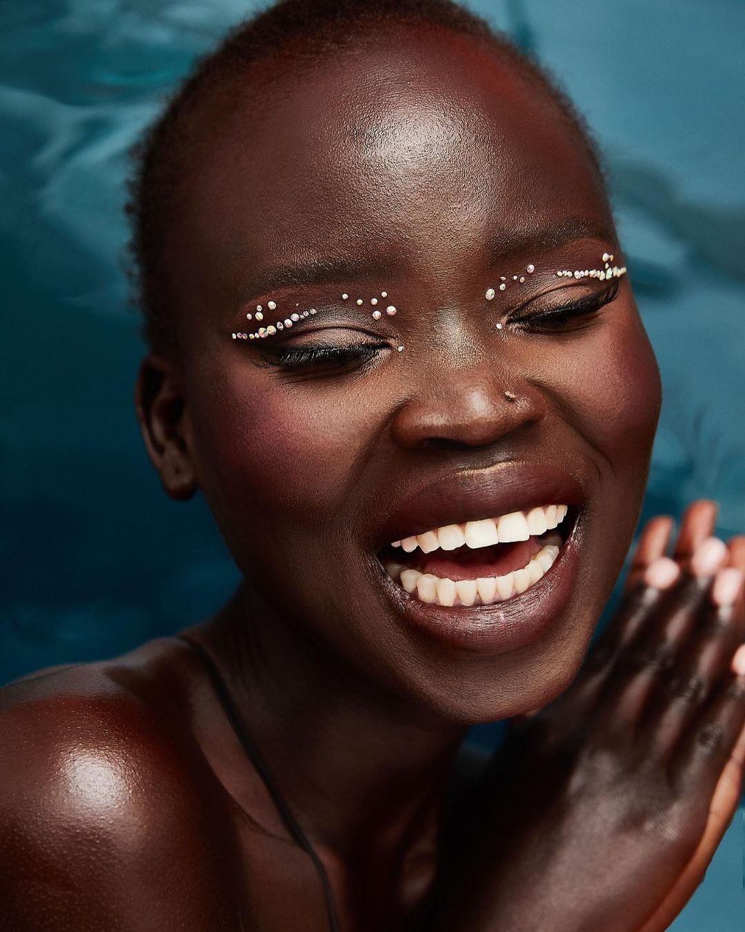 Close-up portrait of a smiling dark-skinned woman with eyes closed, adorned with pearl-like accents around her eyes, against a blue backdrop.