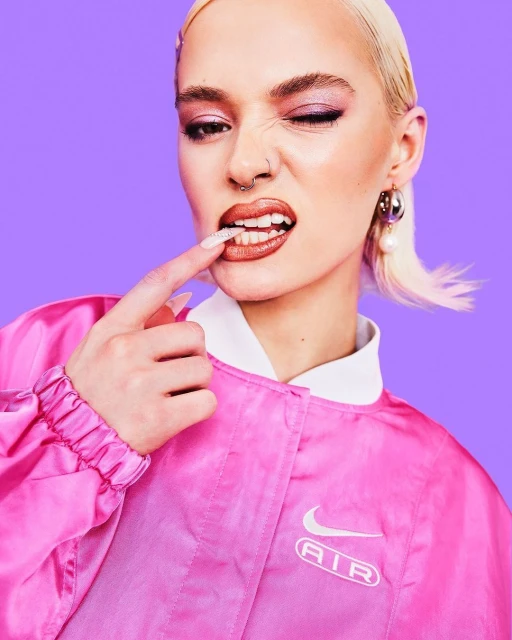 Close-up studio shot of a fair-skinned adult female model winking in a pink jacket, fair hair, light purple background, and light.
