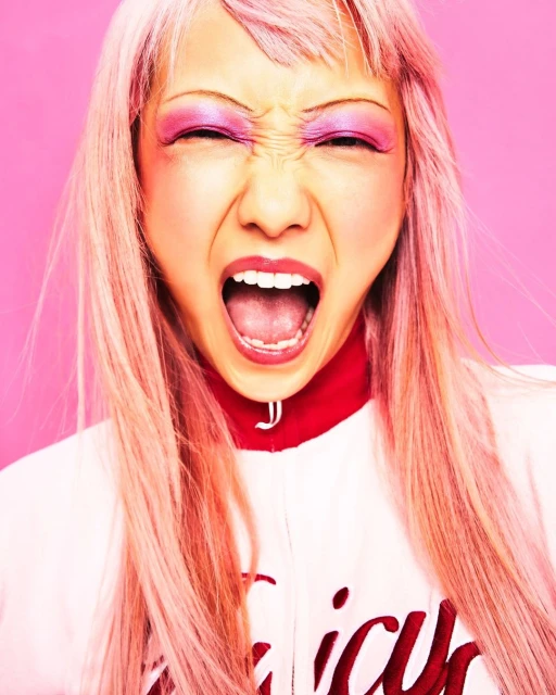 Close-up studio portrait of a woman with pink hair and dramatic makeup, yelling with eyes closed against a pink backdrop. Intense expression.