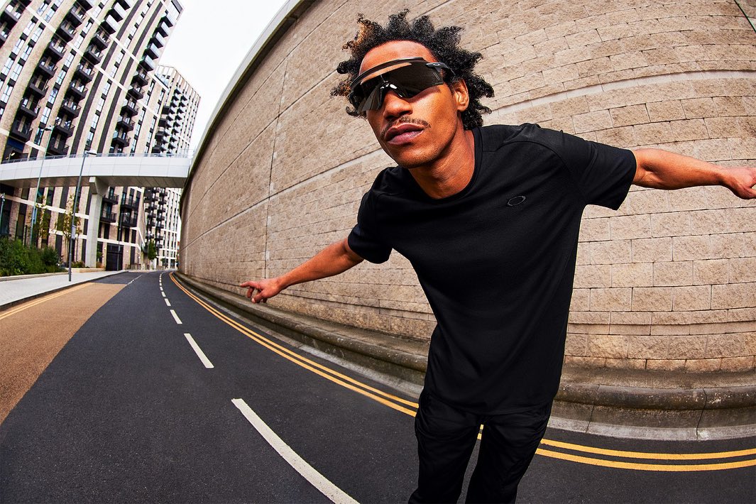 Adult male wearing black sunglasses and black athletic clothing stands on an asphalt road with buildings in the background in an eye-level shot.