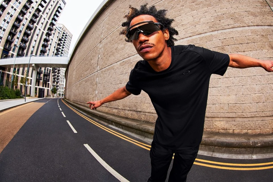 Adult male wearing black sunglasses and black athletic clothing stands on an asphalt road with buildings in the background in an eye-level shot.