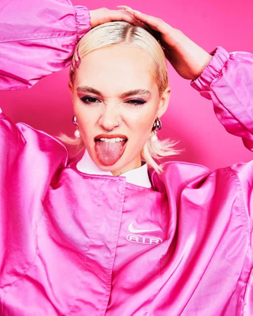 Close-up studio shot of an adult female model with dyed blonde hair sticking out her tongue, wearing a pink jacket with a pink backdrop.