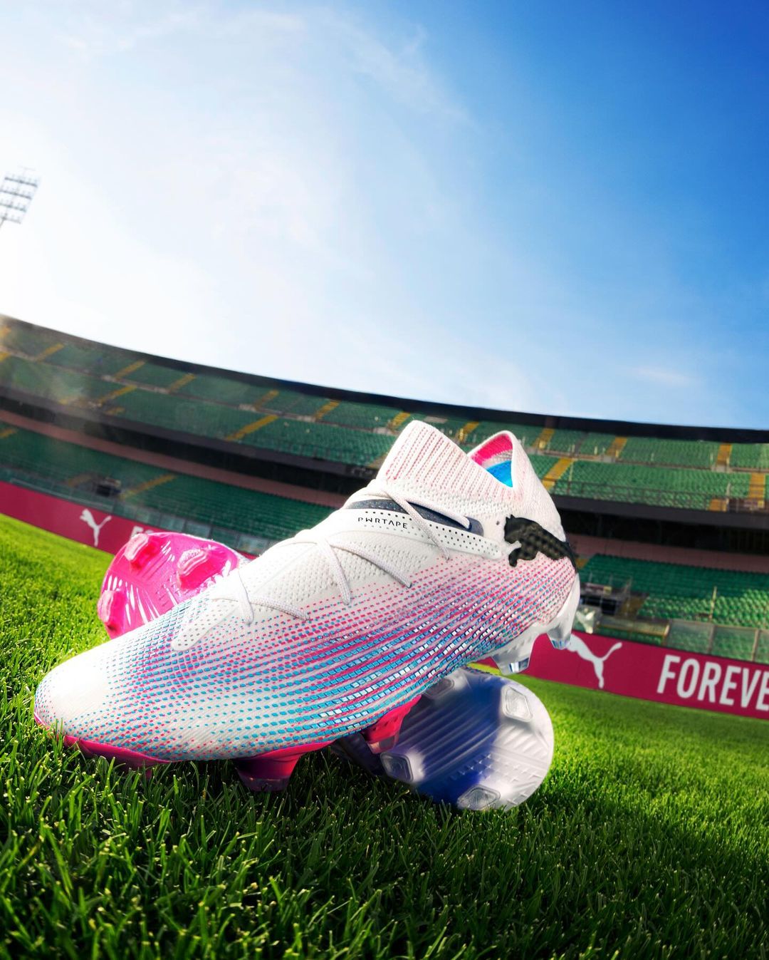 Close-up of a white Puma soccer cleat with pink and blue accents, resting on two soccer balls on green grass with stadium background.