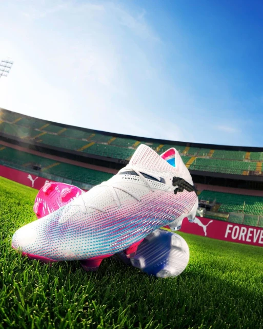 Close-up of a white Puma soccer cleat with pink and blue accents, resting on two soccer balls on green grass with stadium background.