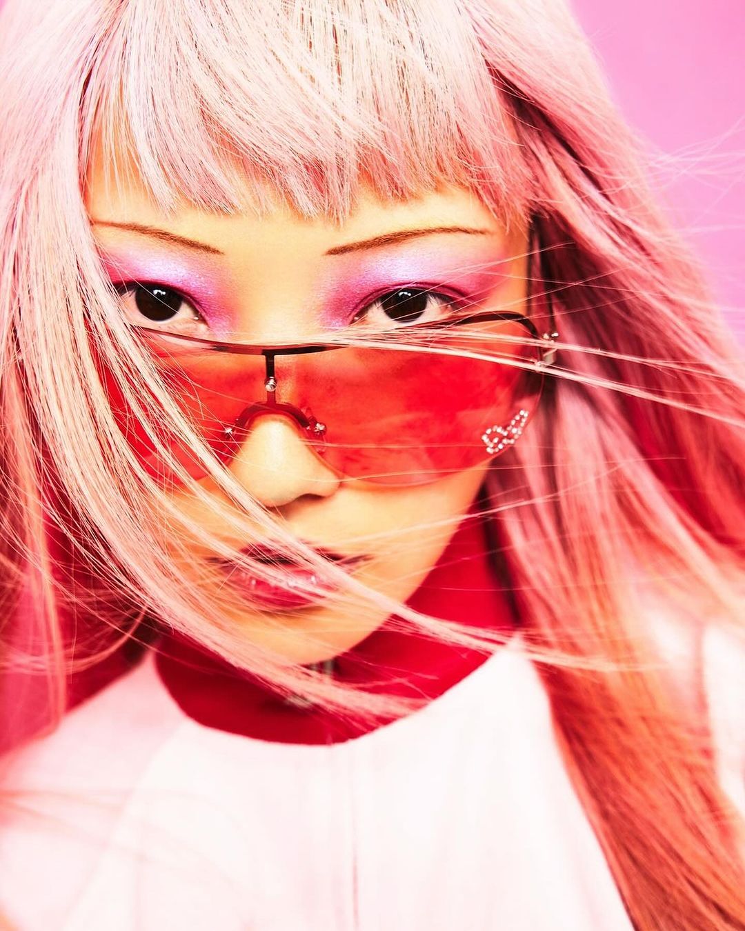 Close-up portrait of a young adult female with pink hair and pink eyeshadow wearing red sunglasses with pink background.