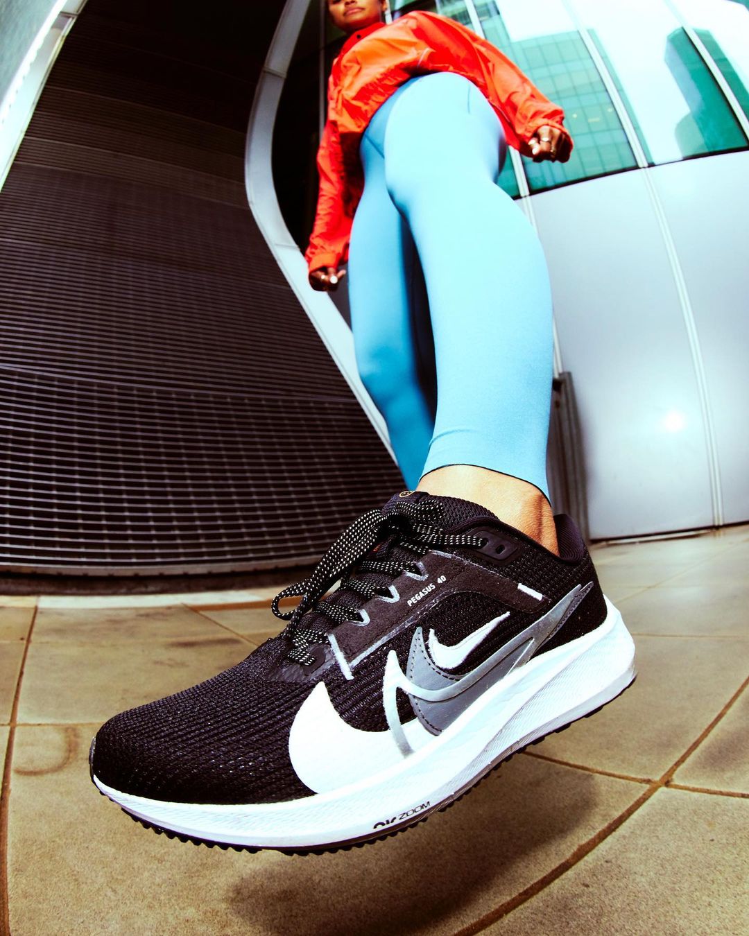 Black Nike running shoe is prominently displayed with an adult in athletic wear posing in an urban environment for a low angle shot.