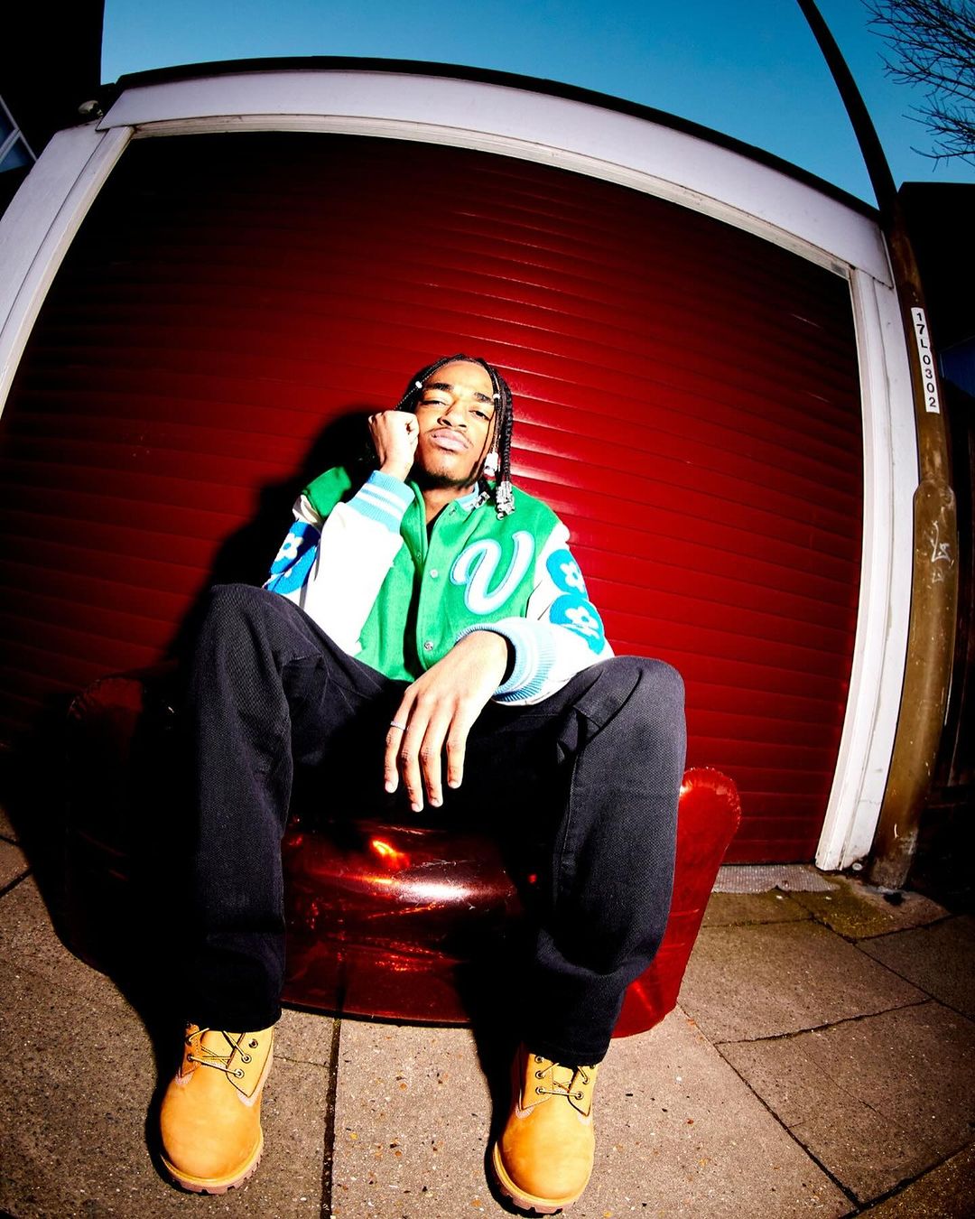 Young man sits in a red chair outdoors in casual fashion, wearing a green jacket, dark jeans, and tan work boots with a garage door backdrop.