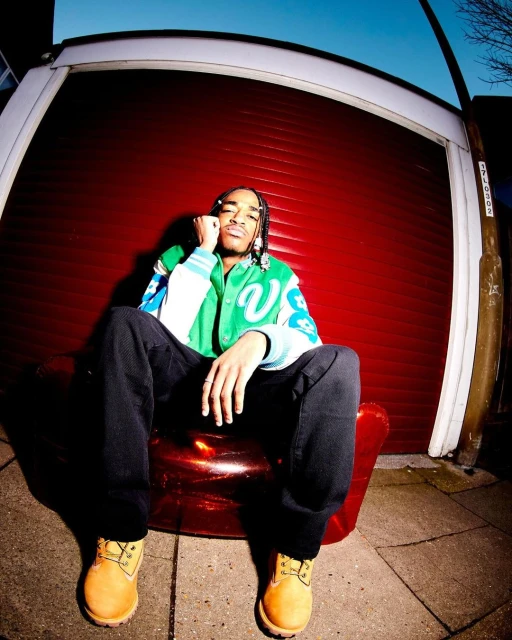 Young man sits in a red chair outdoors in casual fashion, wearing a green jacket, dark jeans, and tan work boots with a garage door backdrop.