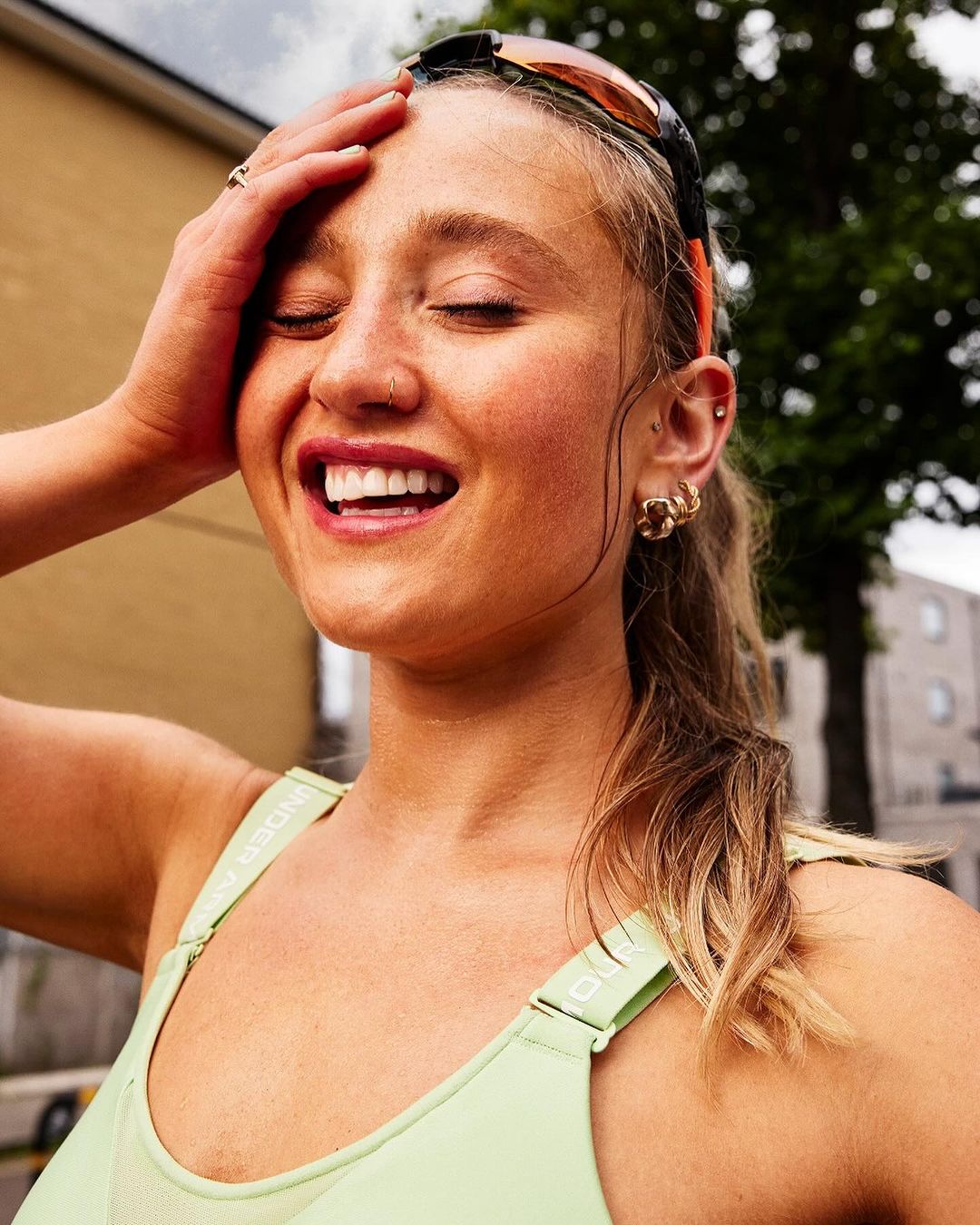 Blonde woman smiles outdoors with hand on head, wearing sporty light green top, buildings and trees in the background.