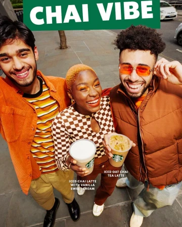 Three smiling adults pose outdoors with iced chai lattes. One wears orange-tinted glasses. Checkerboard shirt, green CHAI VIBE text visible.