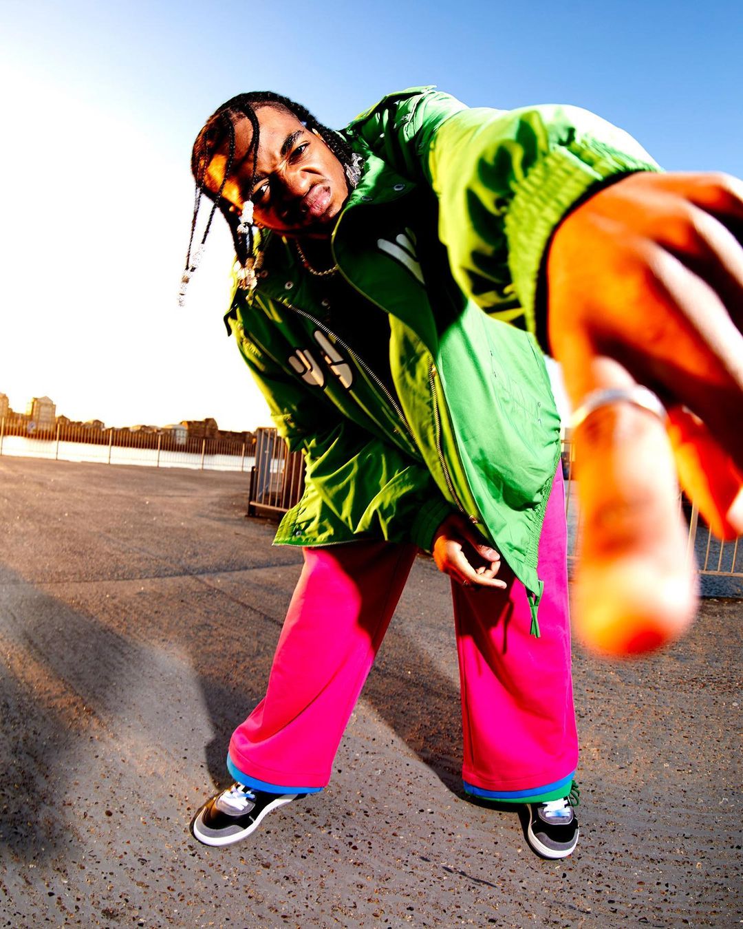Portrait of a young man in a green jacket and pink pants, posing with his arm extended towards the camera in an urban setting.