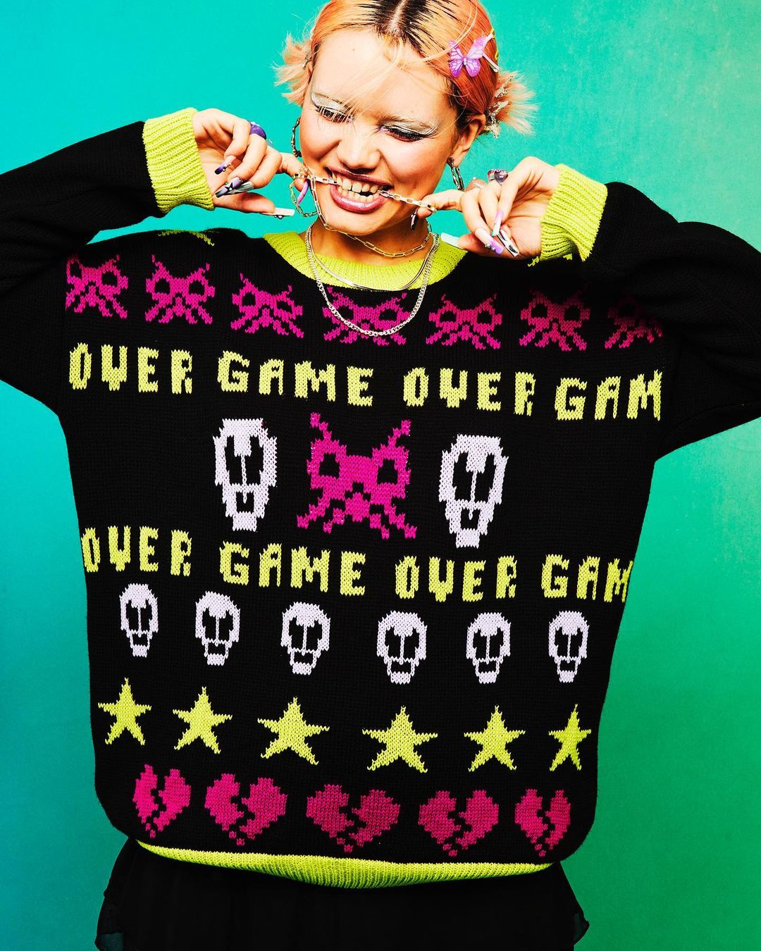 Smiling woman wearing a black knitted sweater with video game pixel art-style graphics, with bright pink and neon yellow details.