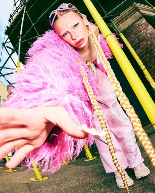 Eye-level outdoor shot of a stylish young woman with blonde braids in a pink fluffy coat, pink pants, and sunglasses, on a city street.