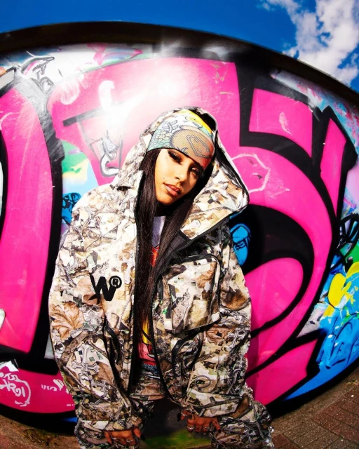 Young woman in camo outerwear poses against graffiti wall under a bright blue sky with some clouds. Stylish urban fashion portrait.