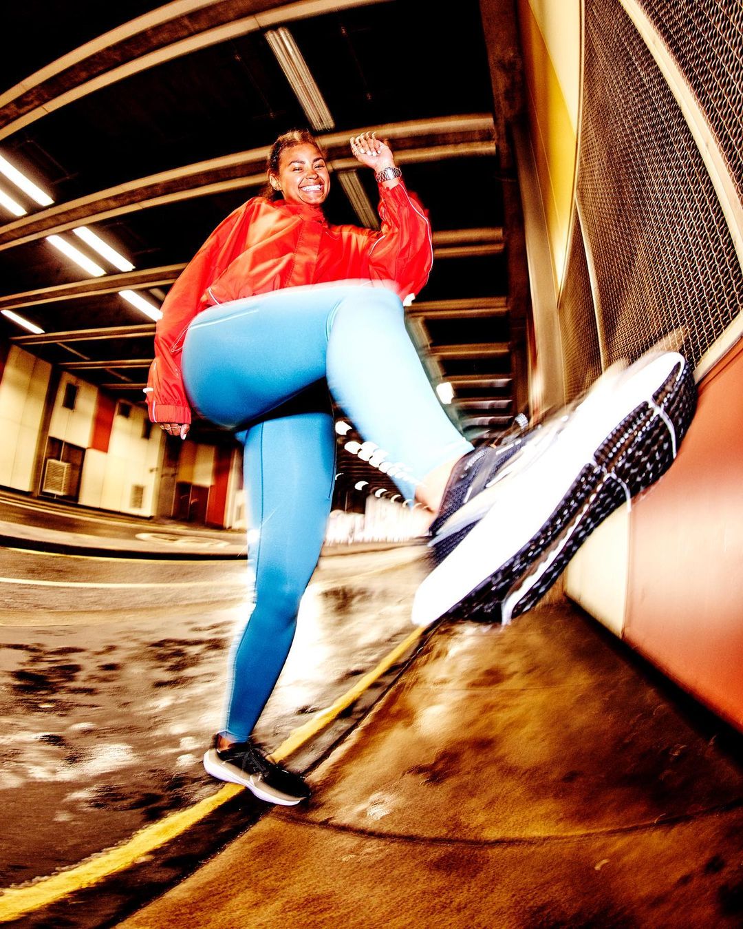 Athletic woman in red jacket and blue leggings strides confidently in a tunnel, showcasing fitness and determination with one foot raised.