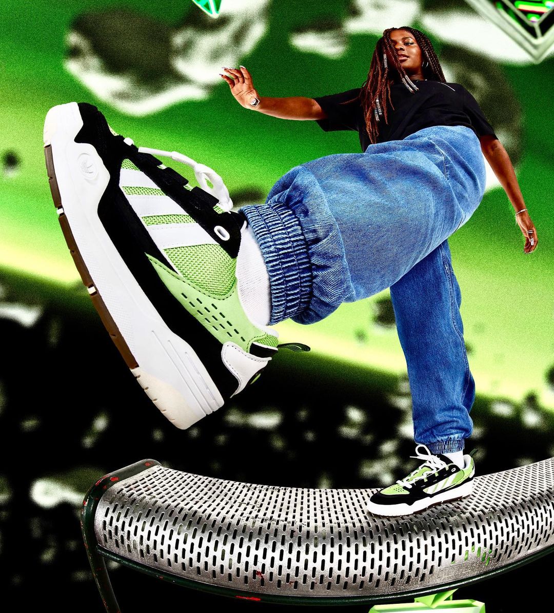 Woman in jeans standing on metal surface, striking pose with green, black, and white sneakers against a green and black backdrop.