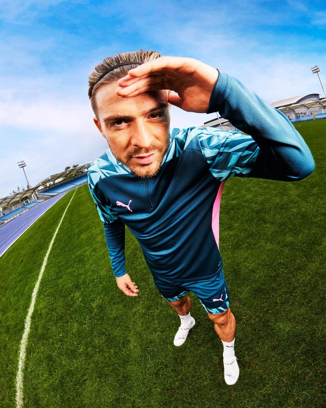Athlete Jack Grealish in sports apparel on a green field with stadium seating. Low angle, eye-level, and wide shot under blue sky.
