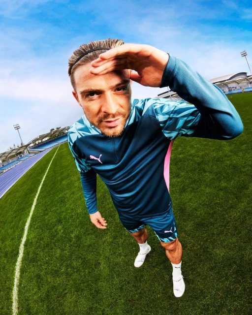 Athlete Jack Grealish in sports apparel on a green field with stadium seating. Low angle, eye-level, and wide shot under blue sky.