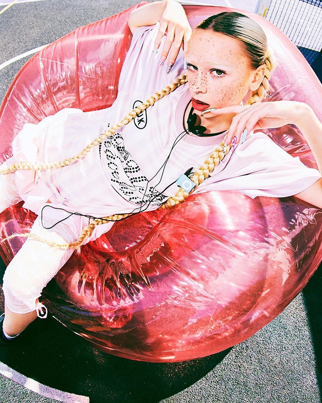 Woman with freckles, braided blonde pigtails, and headphone set wearing light-colored clothing laying on a large, glossy, inflatable pink lip.