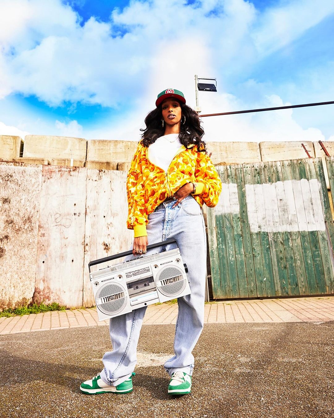 Stylish woman poses with silver boombox outdoors. She is wearing a yellow printed jacket, jeans, baseball cap and green and white sneakers.