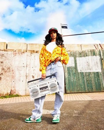 Stylish woman poses with silver boombox outdoors. She is wearing a yellow printed jacket, jeans, baseball cap and green and white sneakers.