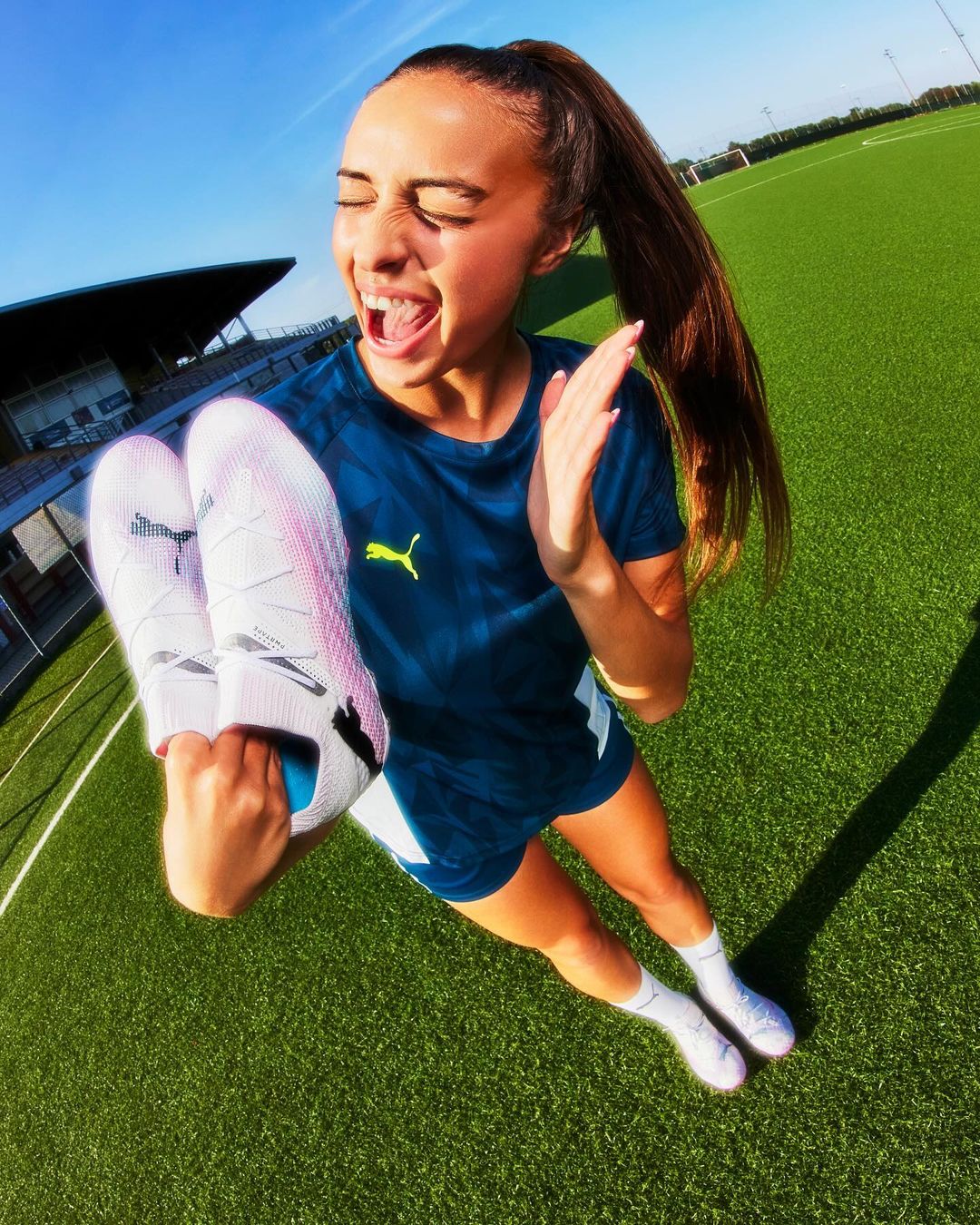 A woman in sportswear holds soccer shoes on a green field, smiling with excitement under a blue sky, with her hair in a high ponytail.