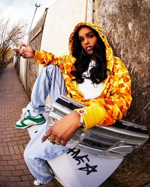 Woman in patterned yellow hoodie sits on silver boombox in alleyway, wearing jeans and sneakers, posing with hand up.