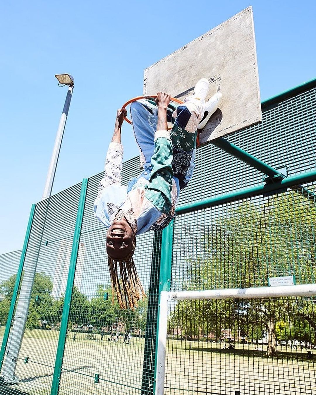 Athletic Black male upside down on an outdoor basketball hoop, wearing a colourful patchwork shirt, blue jeans and white sneakers under a blue sky.