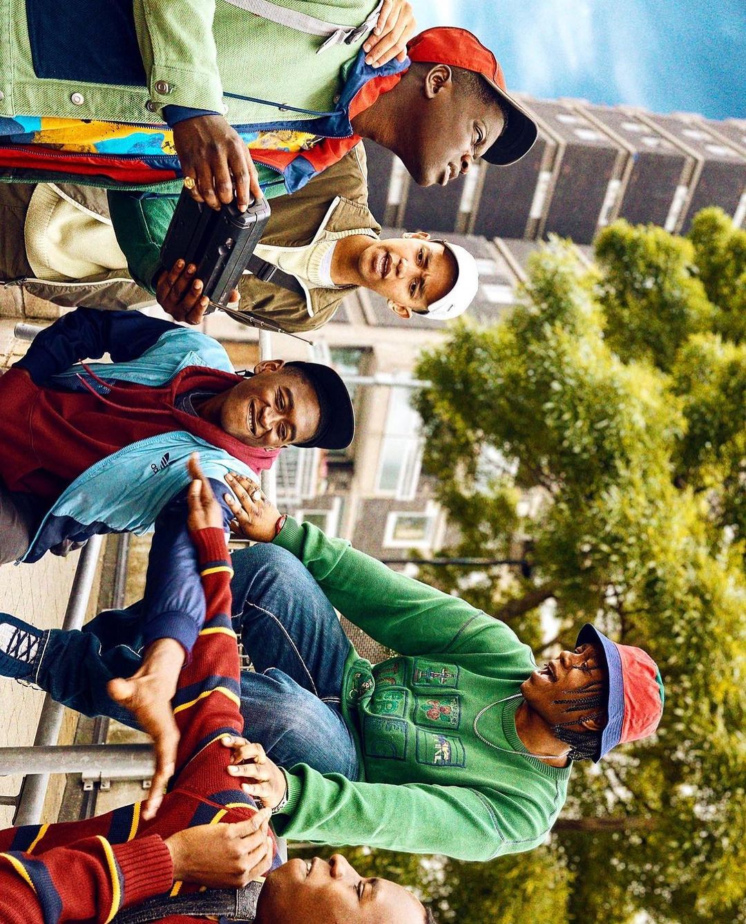 A group of five young adult Black men wearing colourful streetwear stand in an urban setting with trees and a tall building.