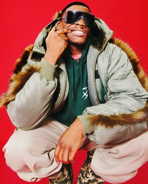 Young man in an olive fur-trimmed hooded jacket and sunglasses squats against a red backdrop, pulling his lip to reveal his teeth.