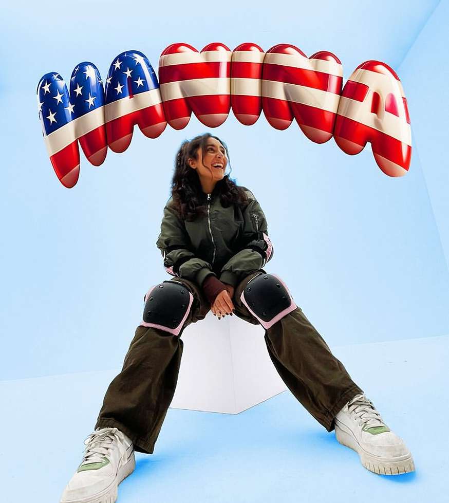 Young woman in an olive bomber jacket and knee pads poses below oversized dental grills decorated with the American flag design.