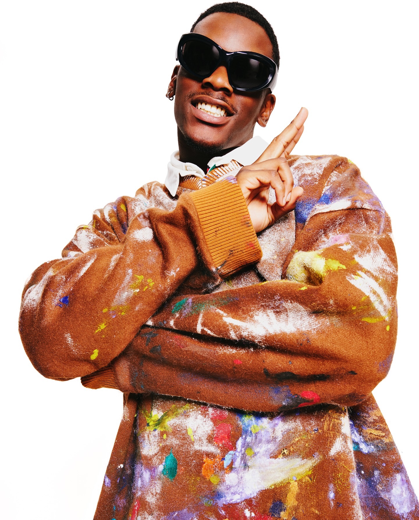 Portrait of a Black male wearing oversized sunglasses and a paint-splattered sweater making a hand gesture against a white background.