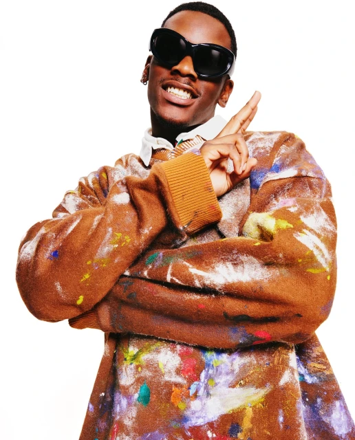 Portrait of a Black male wearing oversized sunglasses and a paint-splattered sweater making a hand gesture against a white background.