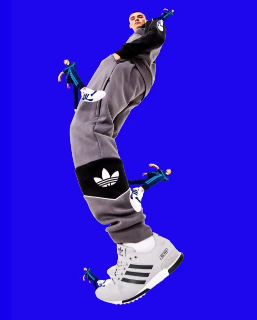 Low angle shot of a man posing in grey Adidas tracksuit, set against a blue background with miniature figures and large Adidas sneakers.
