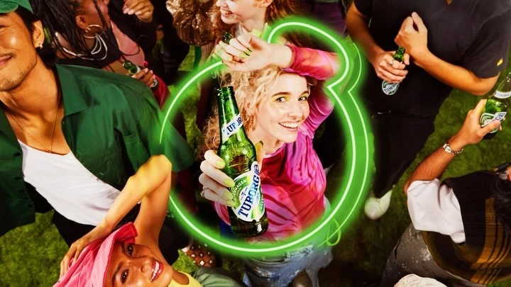 Overhead shot of a smiling woman in a pink tie-dye shirt holding a Tuborg beer bottle, surrounded by other people with beer bottles.