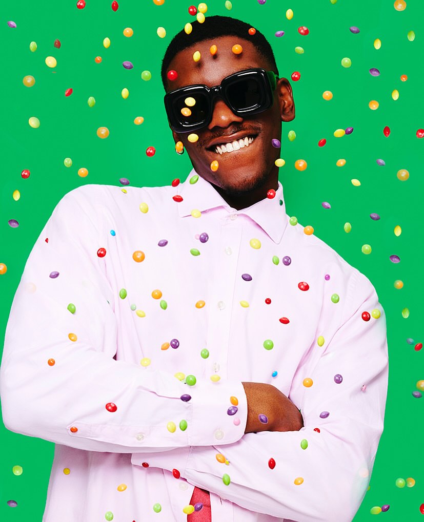 Smiling man in sunglasses and pink shirt poses against a green background with scattered colourful candy pieces around him in a studio setting.