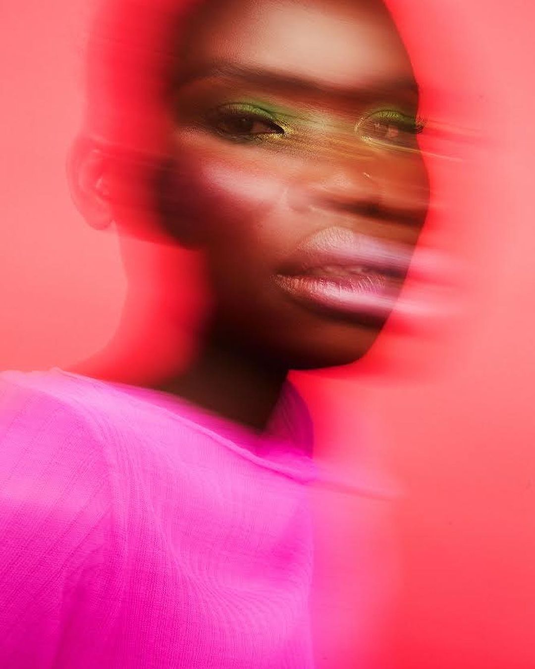 Portrait of a Black model with green eye shadow against a blurred pink background wearing a matching pink top in studio lighting.